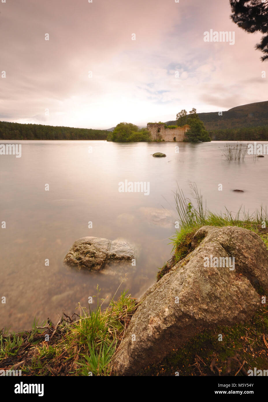 Calmo lago con lunga esposizione, primo piano roccioso, cielo sovrastante e foresta circostante. Loch An Eilein, Aviemore, Scozia Foto Stock