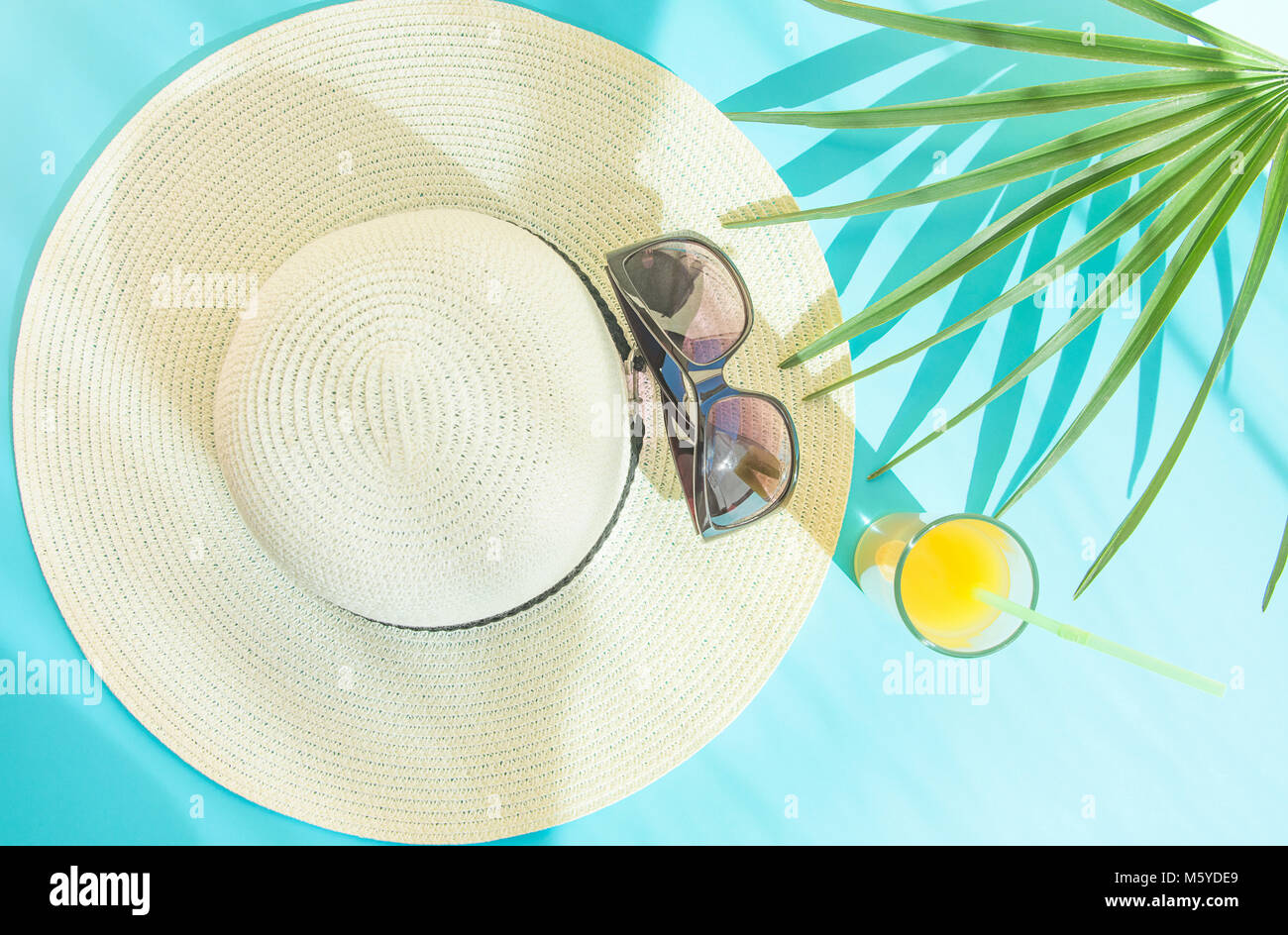 Piatto disposizione laici del cappello di paglia Occhiali da sole di vetro alto con agrumi freschi tropicali Succhi di frutta di foglie di palma su sfondo blu. La luce del sole perdite. Se l'estate Foto Stock