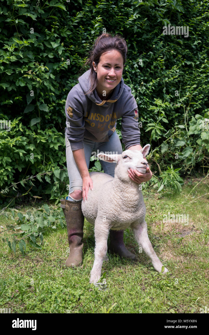 Un giovane agricoltore femmina guarda dopo e feed mentre posa con alcuni agnelli crescente in un campo per la sua azienda. Foto Stock