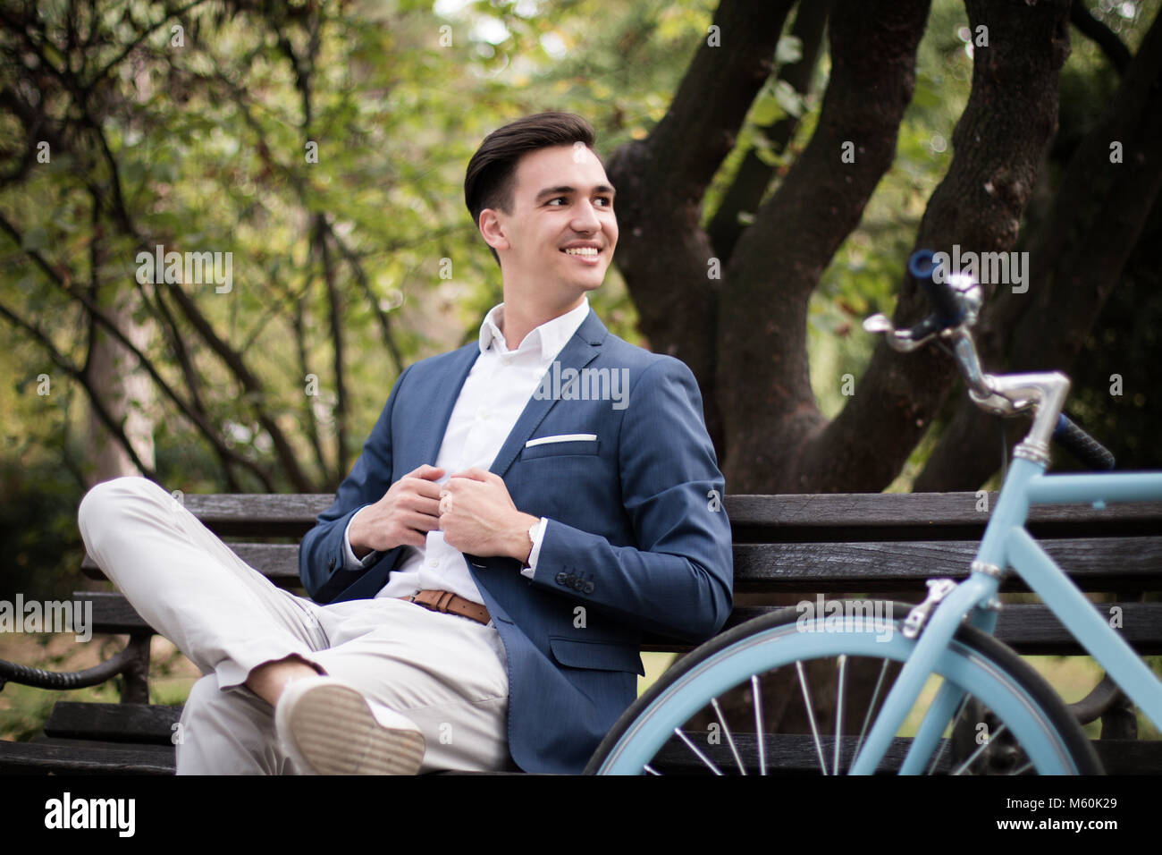 Giovane imprenditore attraente e seduta in attesa su una panchina nel parco con la sua bicicletta accanto a lui Foto Stock
