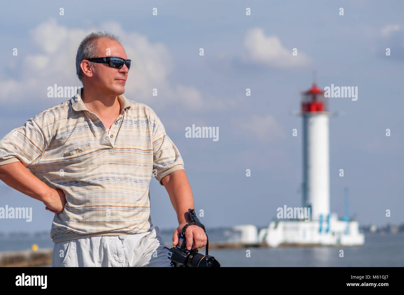 Un uomo di mezza età in una camicia con una manica corta, in occhiali da sole e una fotocamera nera, in un giorno di estate si erge nei pressi di un faro sull'oceano Foto Stock