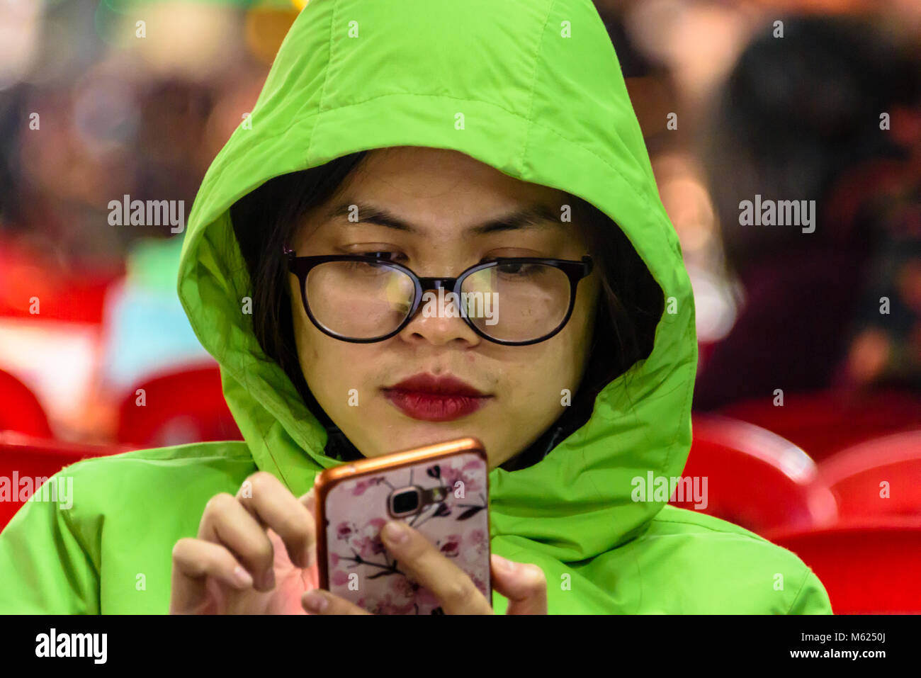 Un vietnamita donna che indossa una felpa con cappuccio verde e rosso rossetto usa il suo smartphone. Foto Stock