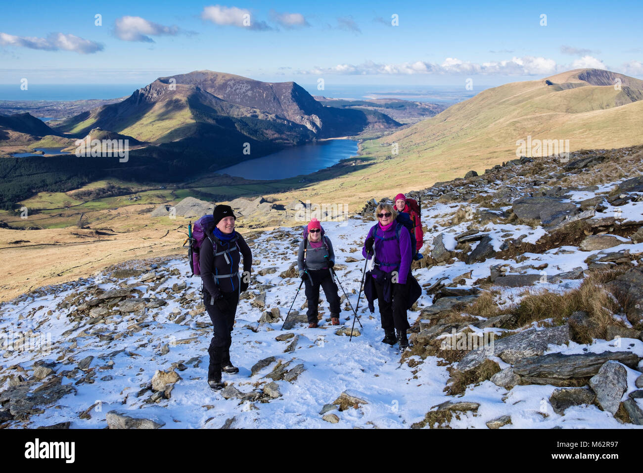 Quattro amici escursionismo su Rhyd Ddu percorso su pendii inferiori di Snowdon con la neve in inverno nel Parco Nazionale di Snowdonia e la vista della costa. Gwynedd, Wales, Regno Unito Foto Stock