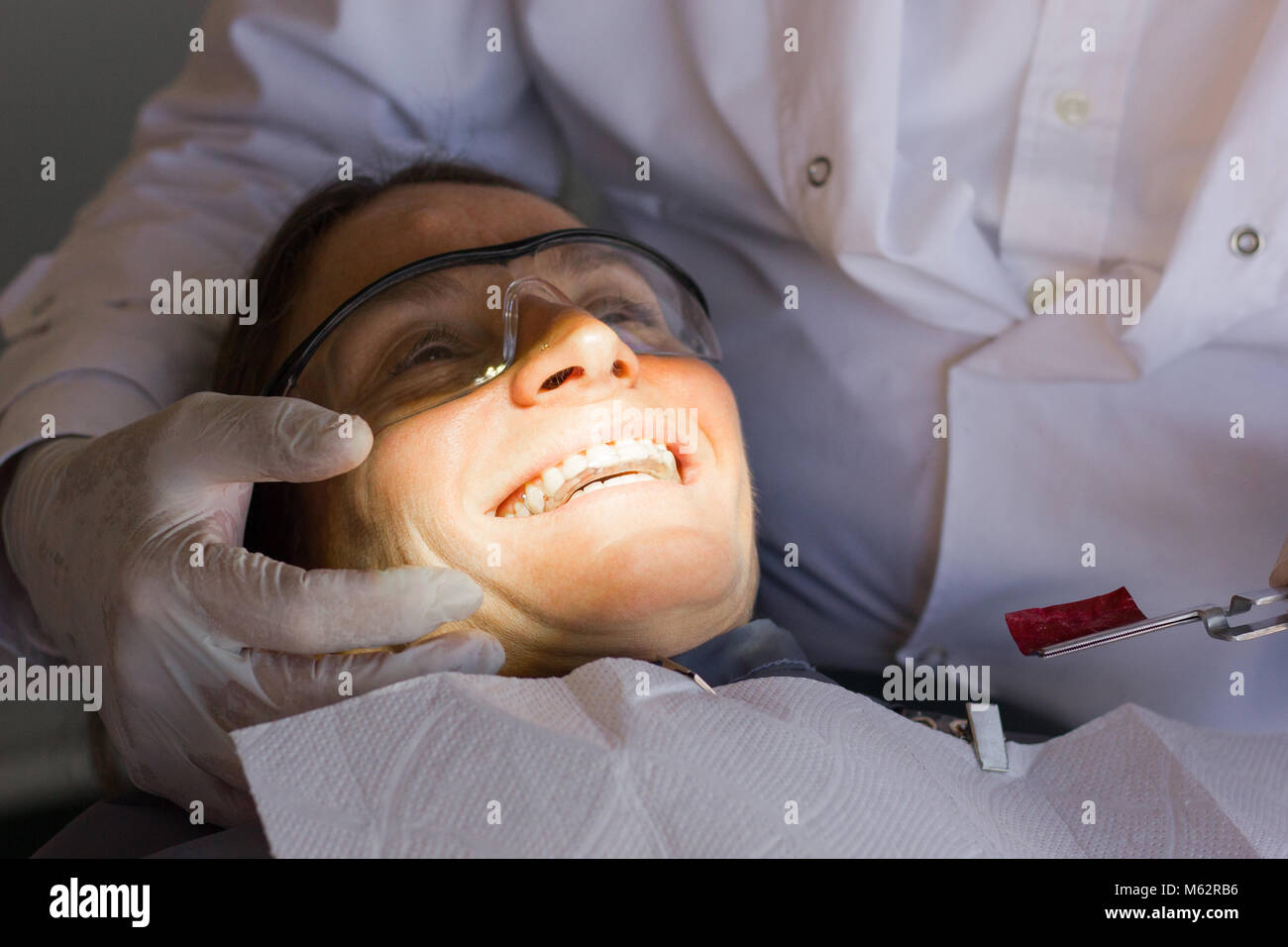 Donna felice con occhiali di sicurezza sorridente con mentoniera a dental clinic dentista mentre tiene il suo volto con dei guanti di protezione in lattice Foto Stock