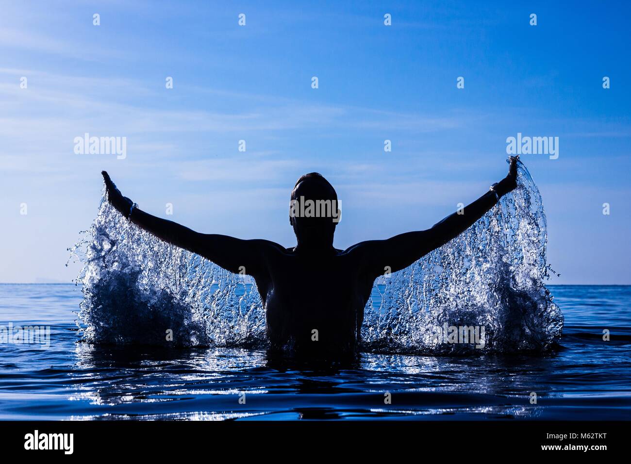 Bloccare il movimento della silhouette di un giovane uomo gli spruzzi di acqua con le braccia aperte in acqua a mystic tramonto dell'isola di Koh Phangan, Thailandia. Filtro blu Foto Stock