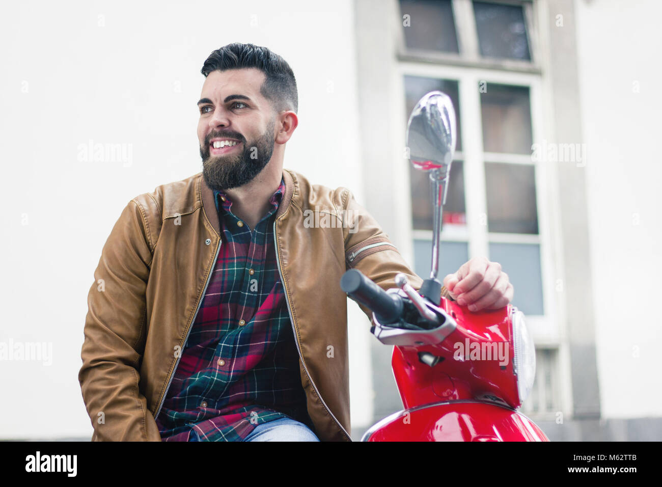 Felice giovane uomo seduto sul motociclo rosso indossando giacca di pelle marrone in città. Hipster con barba folta sorride il classico italiano scooter rétro Foto Stock
