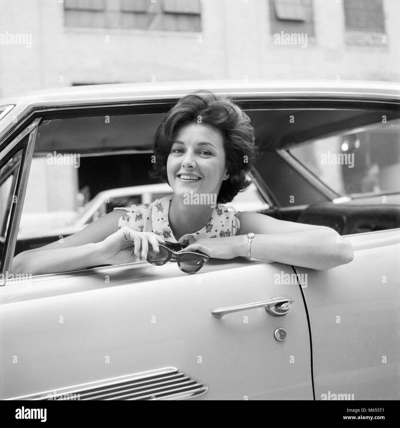 Anni sessanta donna sorridente seduto nel sedile del conducente di vettura appoggiata dalla finestra AZIENDA COPPIA DI OCCHIALI DA SOLE guardando la fotocamera - asp x19197 CAM001 HARS NOSTALGIA il contatto visivo 25-30 anni 30-35 anni BRUNETTE felicità testa e spalle emozione sorriso gioioso automobili VEICOLI DEL DRIVER MID-adulto metà donna adulta B&W in bianco e nero di etnia caucasica guardando la fotocamera in vecchio stile persone Foto Stock