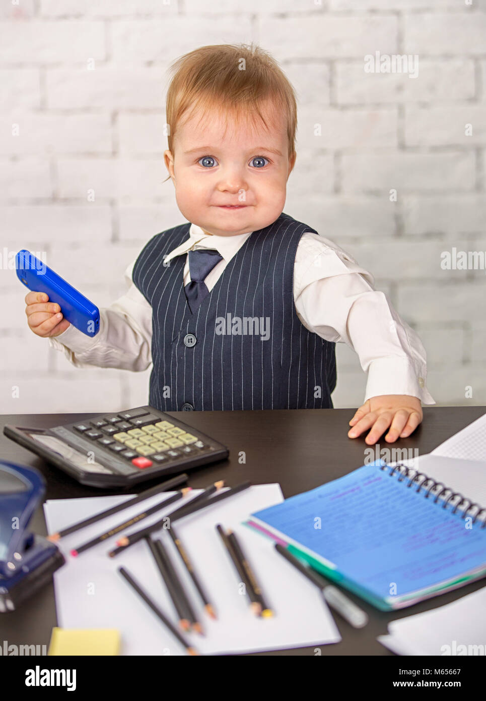 Un simpatico bambino biondo in un business suit è seduto alla scrivania. Broker per neonati con gli elementi di lavoro Foto Stock