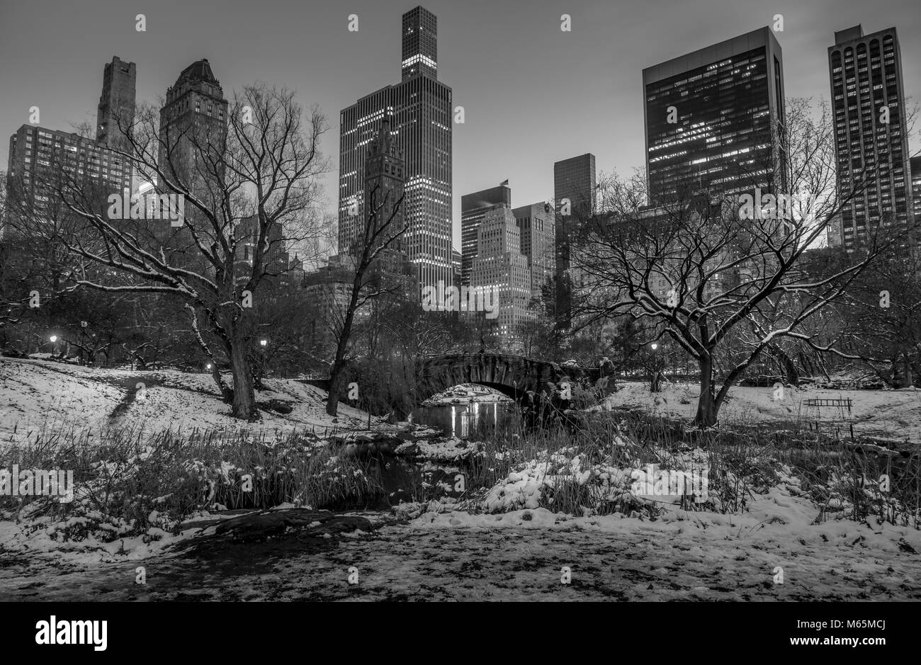 Scena invernale di Gapstow Bridge in Central Park di NY illuminata con alto-aumento in background. Le aree circostanti e alberi coperte con il bianco della neve. Foto Stock