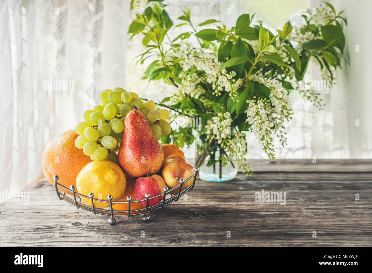 Pera di frutta, succo di pompelmo, limone, pesca sono nel cesto sullo sfondo di un mazzo di piccoli fiori bianchi di bird cherry su un tavolo rustico su un Sun Foto Stock