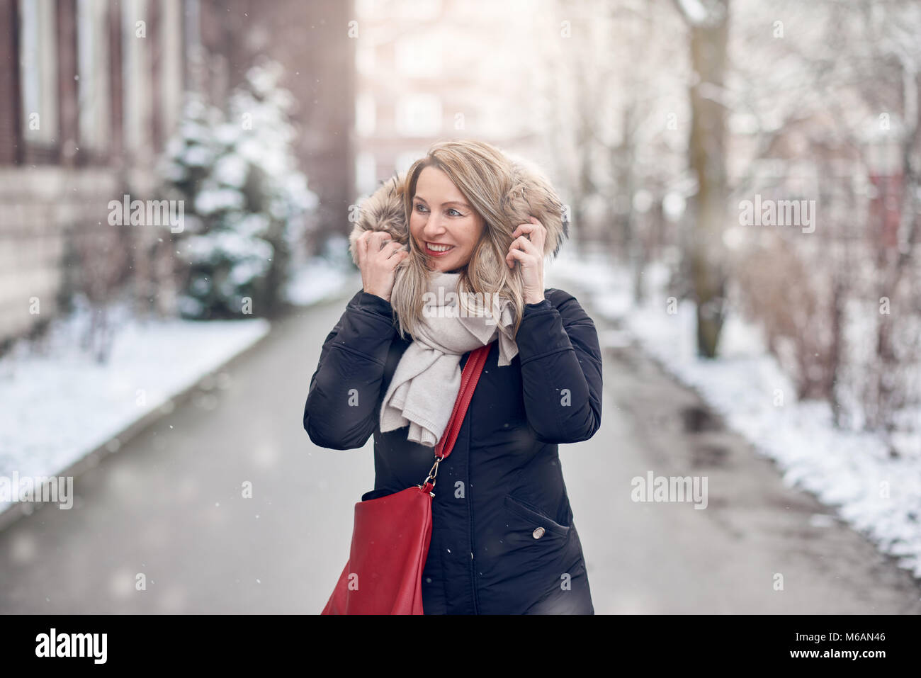 Sorridente ragazza camminare lungo una strada innevata in inverno tenendo premuto il rivestimento in pelliccia per la sua giacca guardando al lato con un sorriso Foto Stock