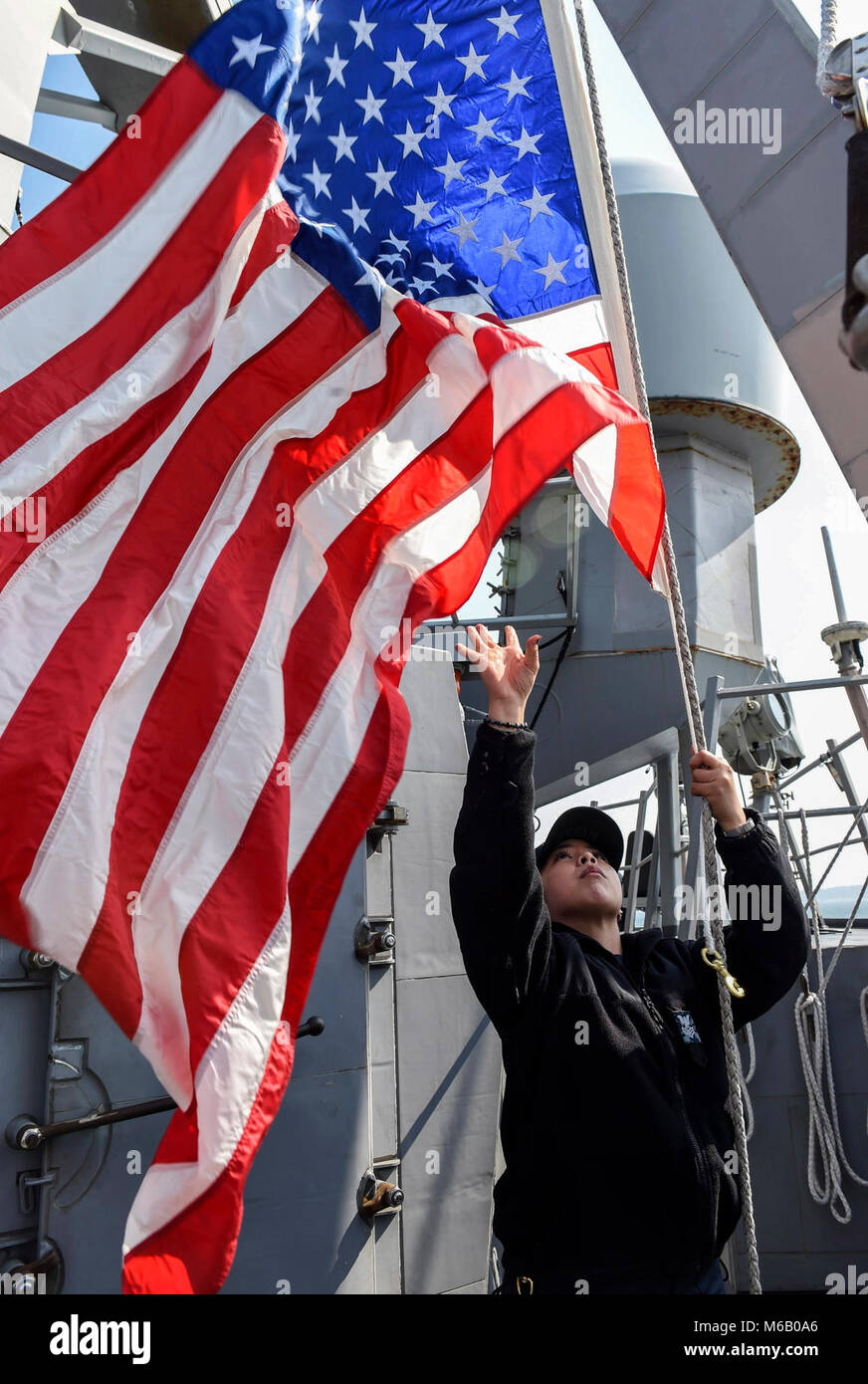 YOKOSUKA, Giappone (feb. 24, 2018) Quartermaster 1a classe Anacristina Nunag, da Brentwood, California, abbassa la bandiera americana durante il mare e i dettagli di ancoraggio a bordo del Arleigh Burke-class guidato-missile destroyer USS Benfold (DDG 65). Benfold è distribuita negli Stati Uniti 7 flotta area di operazioni a sostegno della sicurezza e della stabilità nella regione Indo-Pacifico. (U.S. Navy Foto Stock