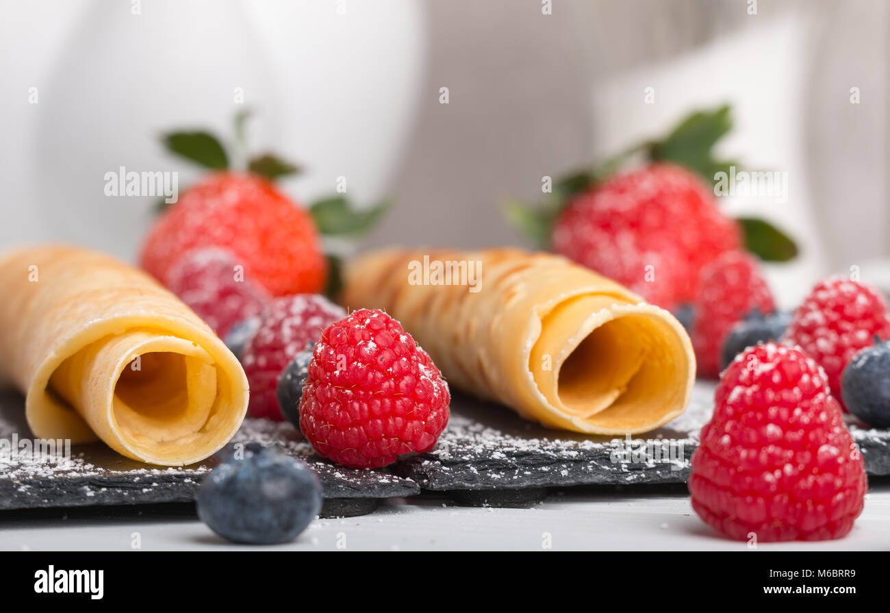 Frittelle di laminati con fresche di fragola e lampone e mirtillo su una pietra nera piastra su un bianco tavolo in legno con il miele e lo zucchero bianco e Foto Stock