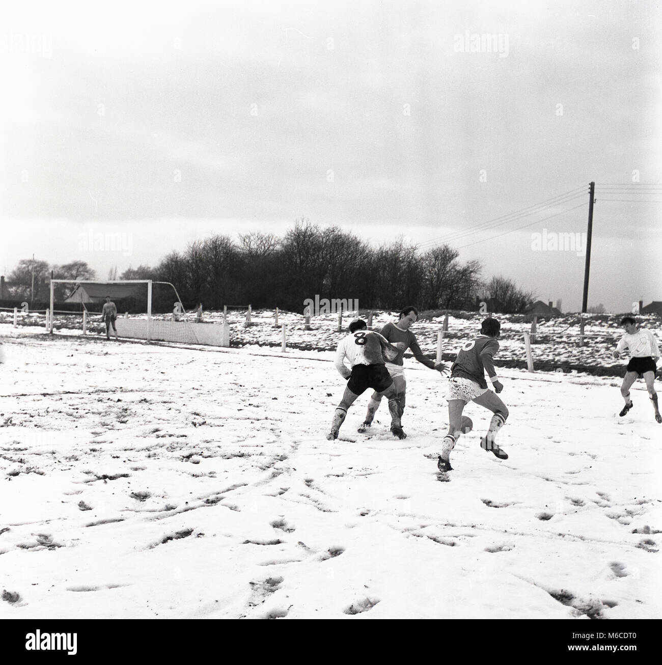 Anni sessanta, storico inverno e calciatori dilettanti giocando una partita su una coperta di neve passo, Inghilterra, Regno Unito. Foto Stock