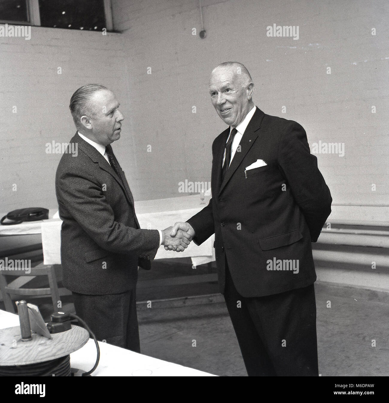 Anni sessanta, England, Regno Unito, immagine mostra due ben vestito uomini che indossano tute in piedi agitando mani. Una stretta di mano ferma - con la loro mano destra- da un uomo ad un altro è un tradizionale sistema britannico di scambio culturale o rituale e in questo caso, è associato con il messaggio 'tutti i migliori' o 'auguri sul vostro reitrement'. Foto Stock