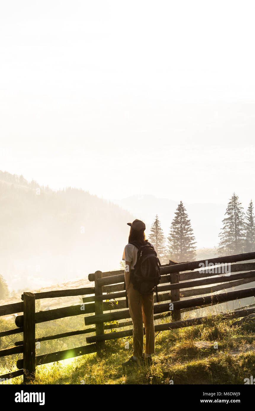 I giovani backpacker ragazza turistico sorge dal recinto e guarda alle colline con foreste e la nebbia di sunrise Foto Stock