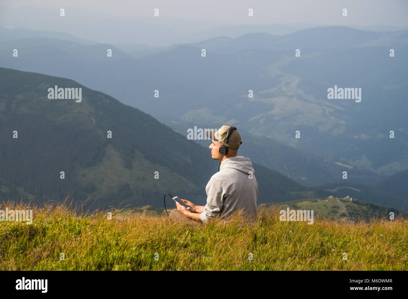 Giovane maschio persona gode di musica in cuffia dal telefono cellulare su un prato di montagna Foto Stock