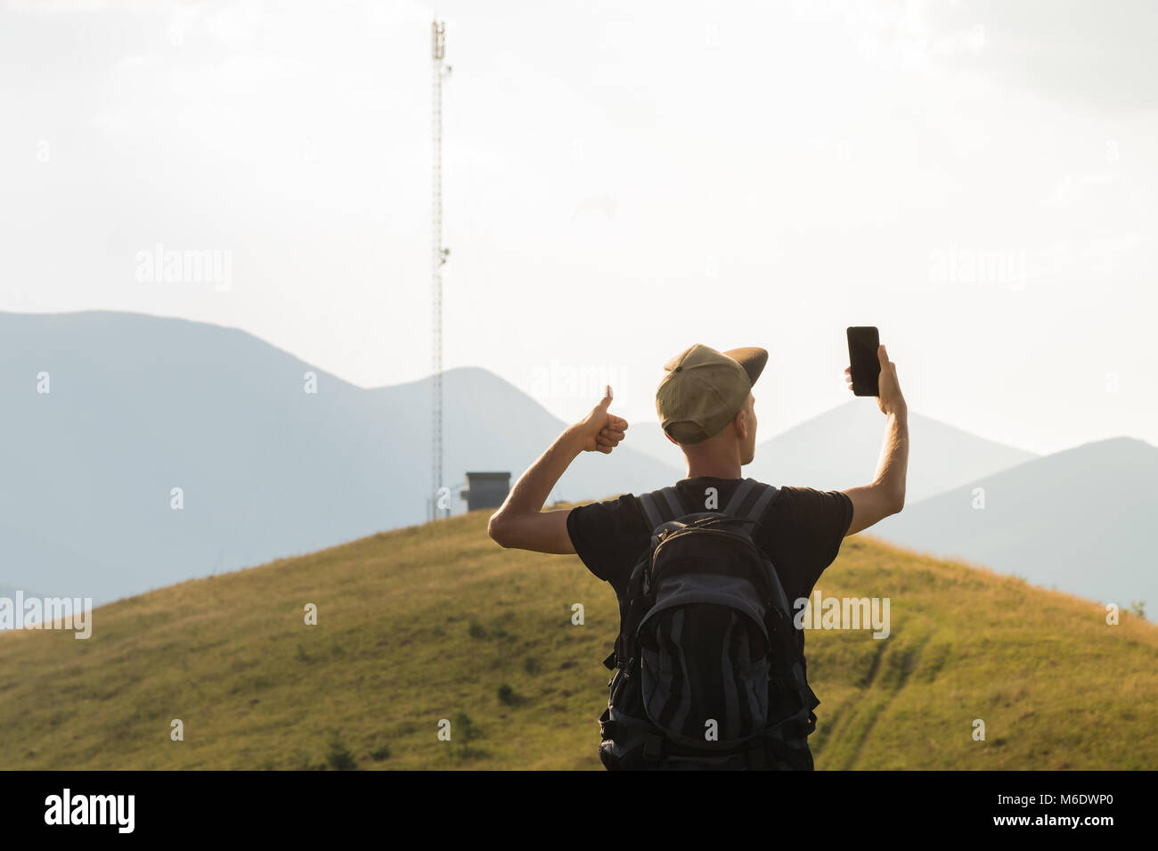 Giovane uomo in vacanza in montagna alza il pollice in alto e detiene il telefono cellulare vicino alla cella della torre di rete Foto Stock