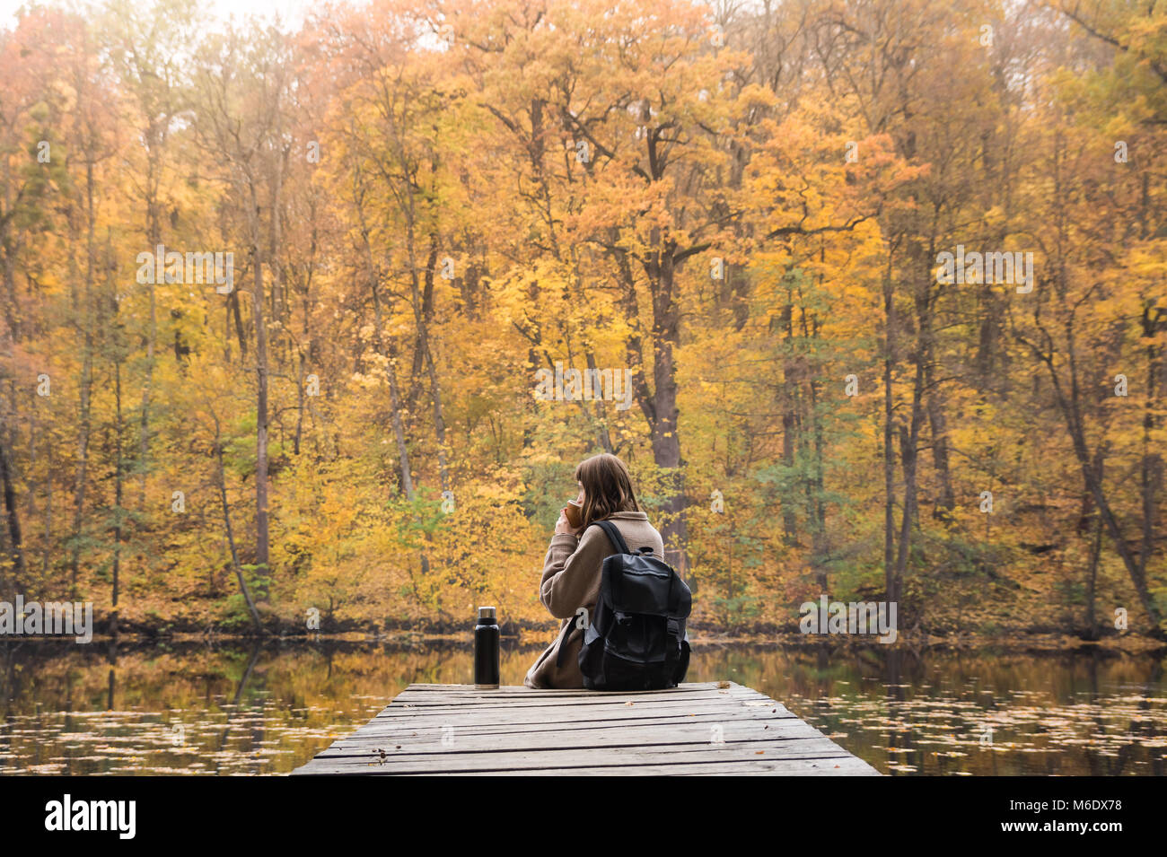 Giovane persona di sesso femminile di escursioni presso il parco di natura si appoggia a riverbank su oro giornata autunnale e gode di una bella ottobre scenario avente una bevanda calda da thermos Foto Stock