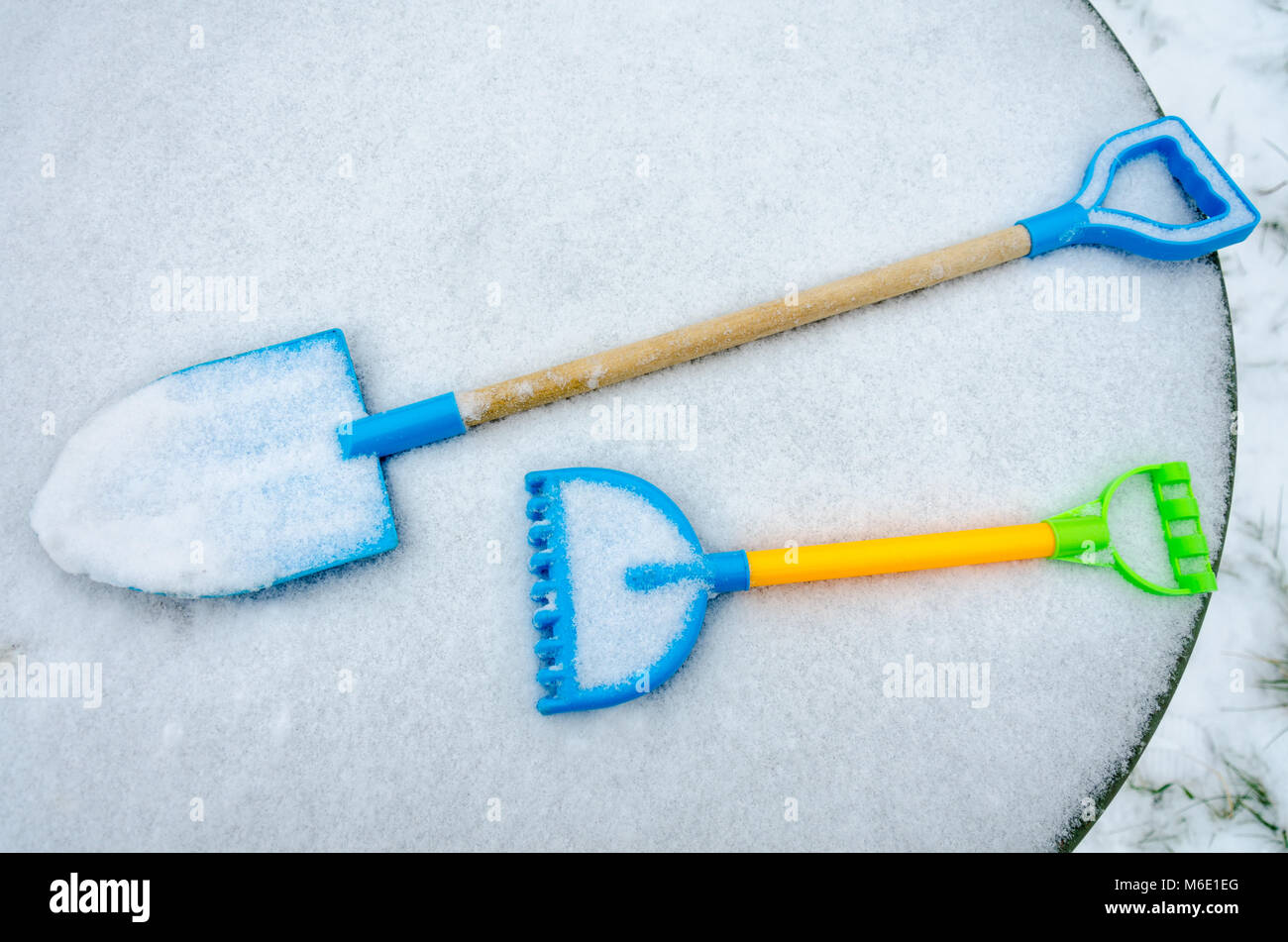 Un bambino giocattolo vanga e rastrello sono coperti di neve che è stato lasciato fuori su un tavolo da giardino prima ha nevicato. Foto Stock