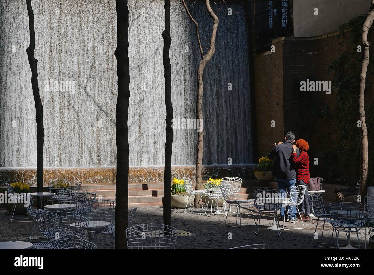 New York, NY, STATI UNITI D'AMERICA: l uomo e la donna a guardare la cascata nella tasca Paley Park, Midtown Manhattan, amid Tulipani gialli e tavoli e sedie bianchi. Foto Stock