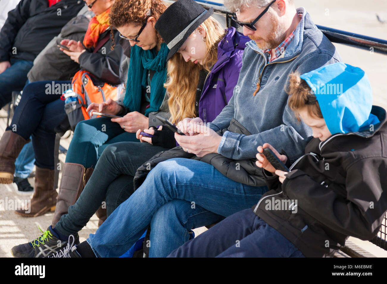 Famiglia seduta su una panchina, tutti dei quali sono simultaneamente guardando il loro telefono cellulare / Dispositivo / dispositivi, scrivere messaggi o navigare utilizzando / web / internet. Foto Stock