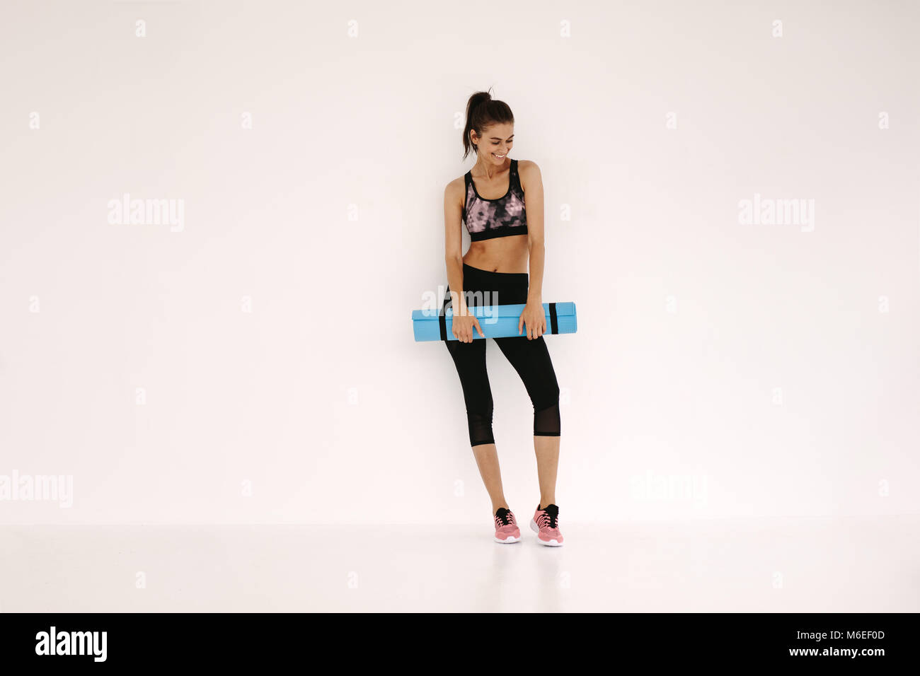 Sportive in piedi con materassino yoga in fitness studio. Un sano modello femminile in piedi su sfondo bianco con materassino yoga dopo allenamento. Foto Stock