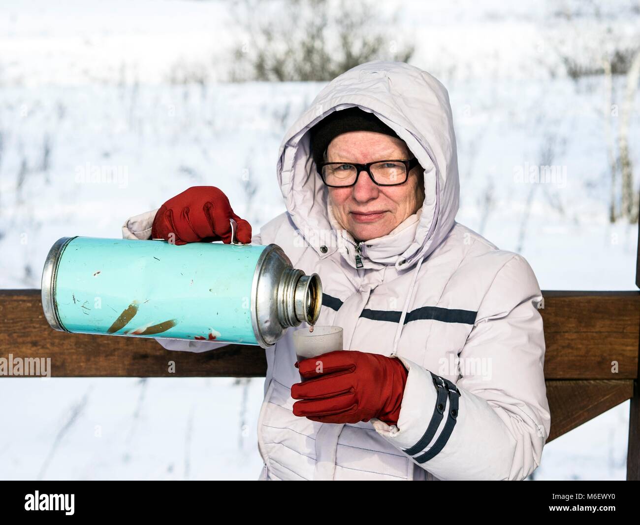 Caucasian donna di mezza età versa il tè da thermos. seduta su una panchina in un gazebo sullo sfondo di una coperta di neve campo e foresta Foto Stock