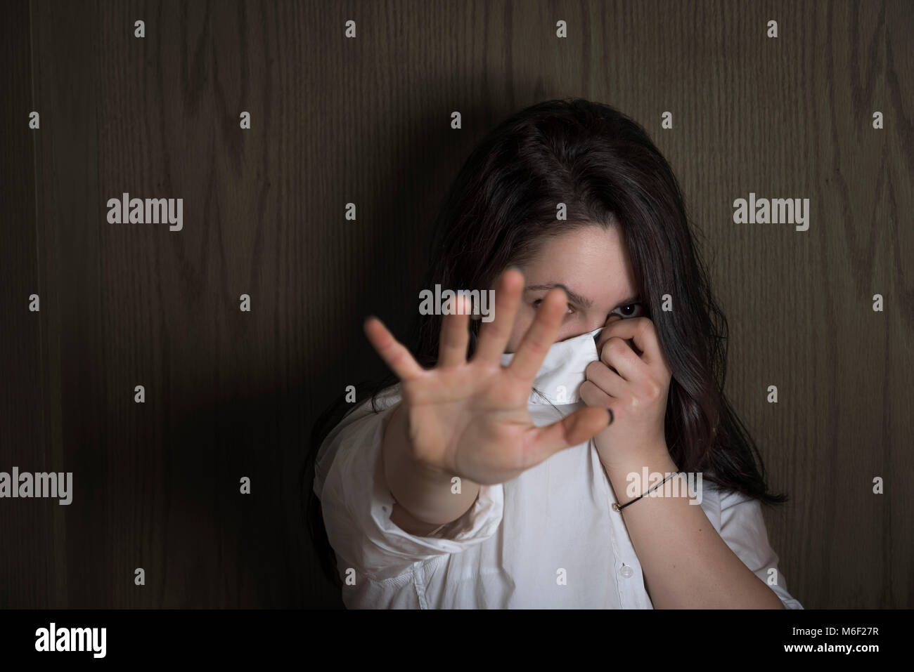 Giovane donna nasconde il suo volto con la sua camicia mentre si estende la sua mano verso la telecamera in modo da non essere visto su un sfondo di legno, in ombre. Foto Stock
