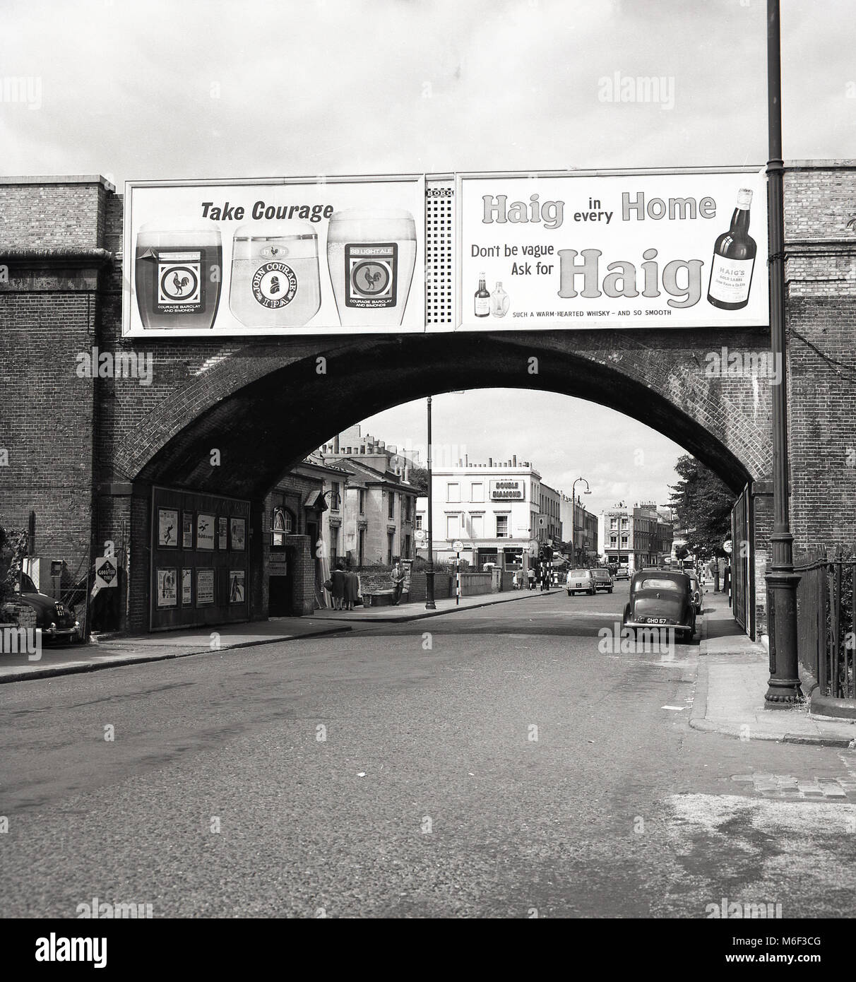 Anni sessanta, foto storiche di una tranquilla Kentish Town Road, London, NW1, mostrando grandi pannelli pubblicitari per le grandi marche di bevande del giorno, coraggio birra e whisky Haig su un ponte ferroviario. In lontananza, un segno per un altro ben noto la birra del giorno, 'Double diamante' da Ind Coope a Moreton Arms pub. Foto Stock