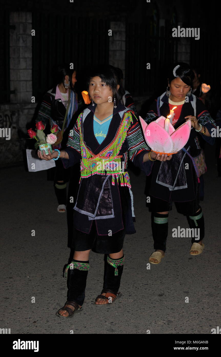 Giovani felici e solenne Hmong ragazze vestiti in abiti tradizionali tenendo candele e fiori celebrare la Vergine Maria a Sapa chiesa di pietra di notte. Foto Stock