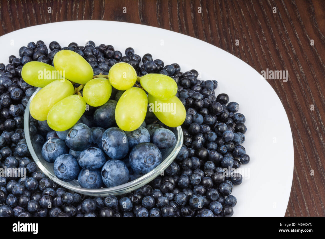 I mirtilli e il grappolo di uva sulla tavola. Vaccinium myrtillus. Close-up di piastra bianca con fresche bacche e ciotola di vetro su sfondo di legno. Foto Stock