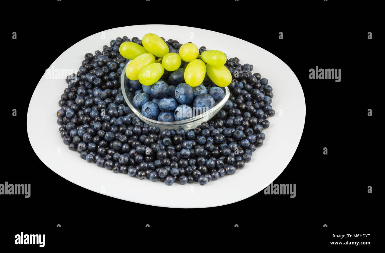 Organici di mirtilli e grappolo di uva. Vaccinium myrtillus. Grandi e piccoli frutti a bacca bianca sulla piastra e ciotola di vetro. Isolato su sfondo nero. Foto Stock