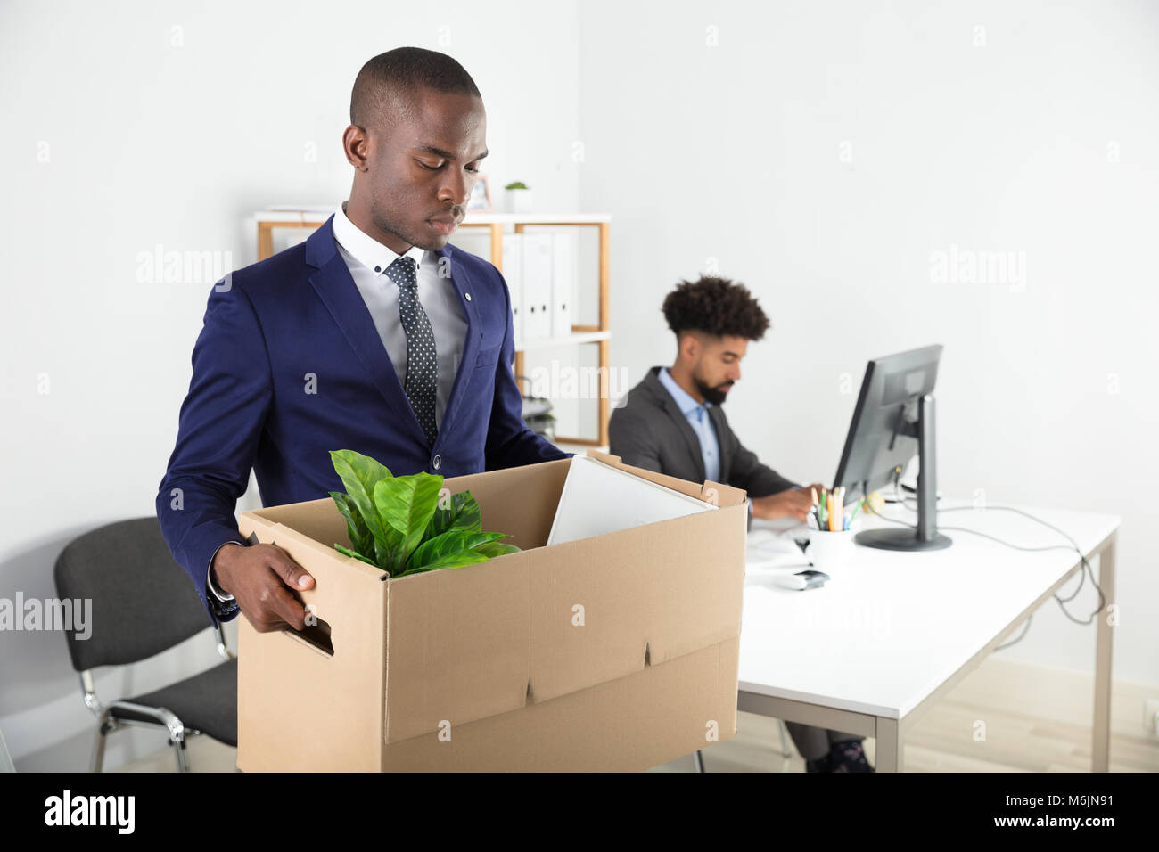 Imprenditore si sposti fuori con una scatola di cartone da ufficio con un dipendente che lavora presso la scrivania Foto Stock