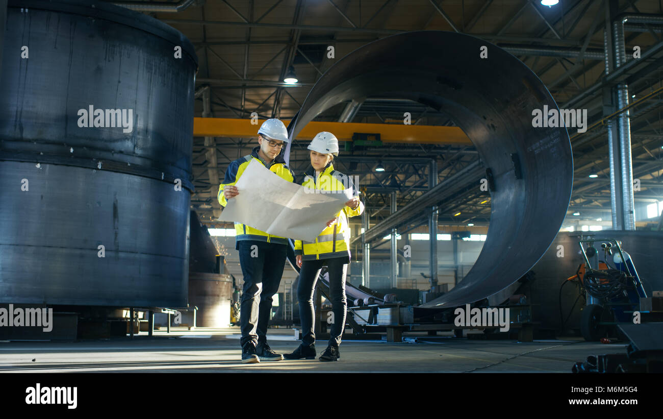 Maschio e femmina ingegneri industriali guardare al progetto Blueprint mentre Surround permanente dalle parti della pipeline nel mezzo di enorme industria pesante Foto Stock