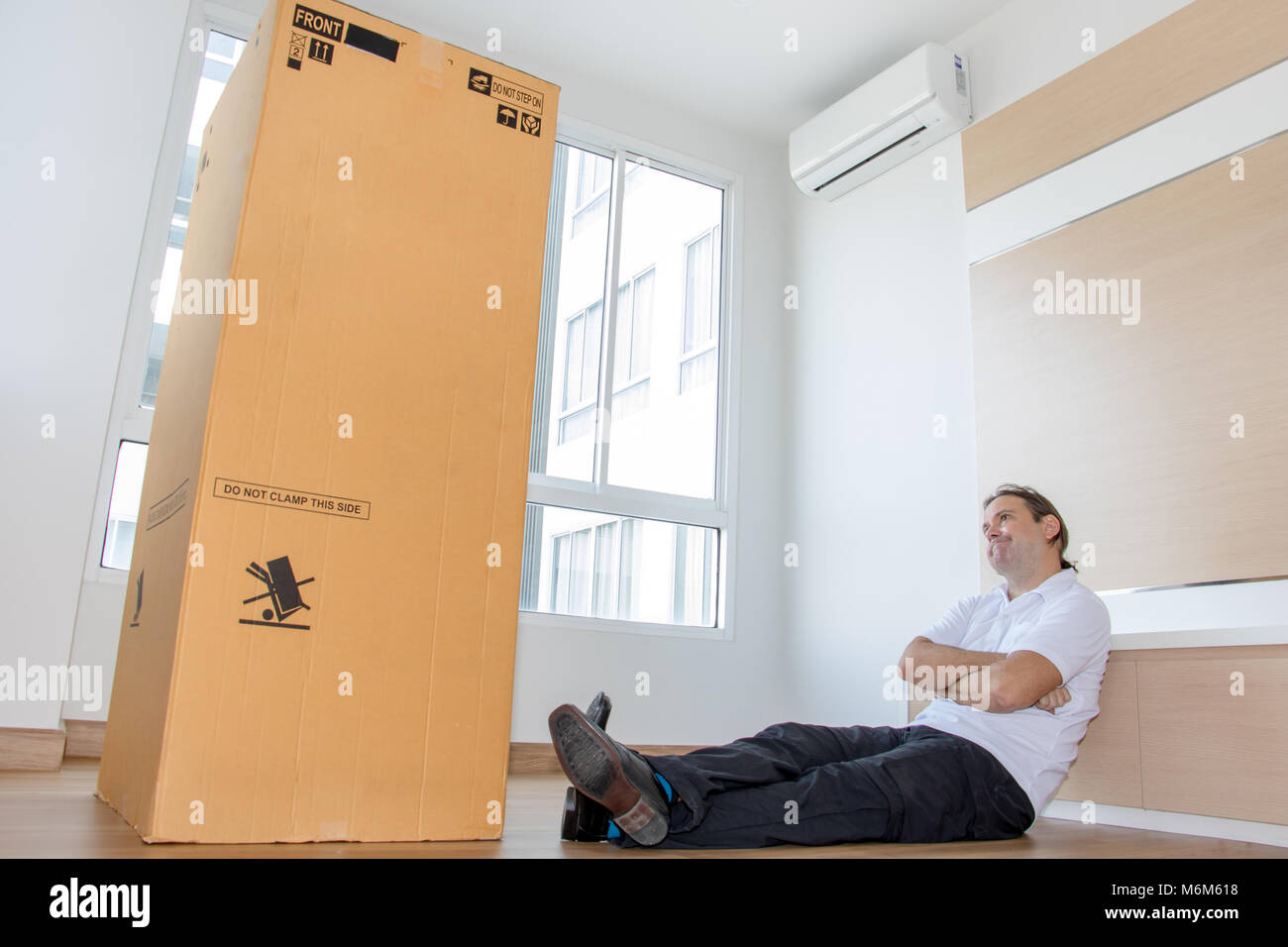 Uomo seduto sul pavimento in un appartamento vuoto guardando una grande scatola di cartone. L uomo è il pensiero del grande pacchetto nel nuovo appartamento. Foto Stock
