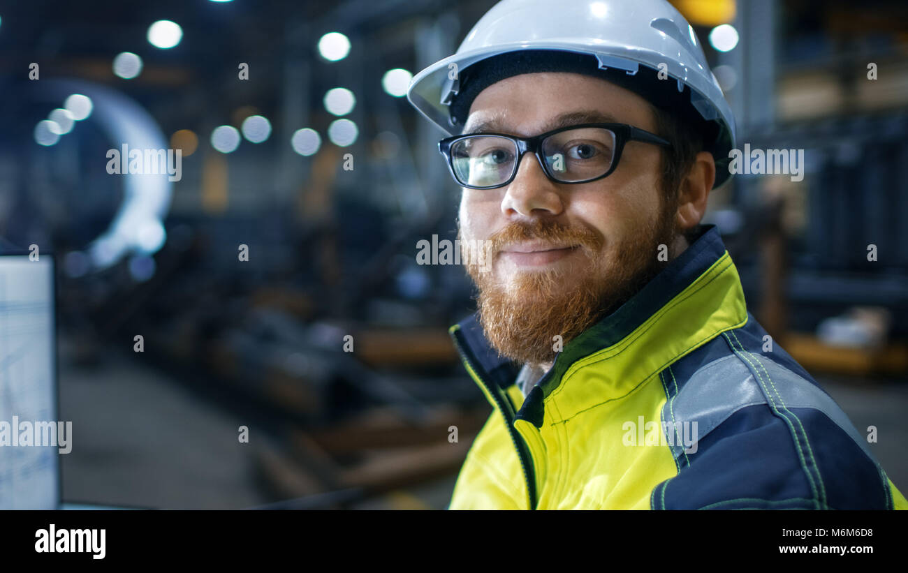 Ingegnere Industriale indossare elmetto, giubbotto di sicurezza e bicchieri sorrisi sulla fotocamera. Egli lavora in grande industria pesante fabbrica. Foto Stock