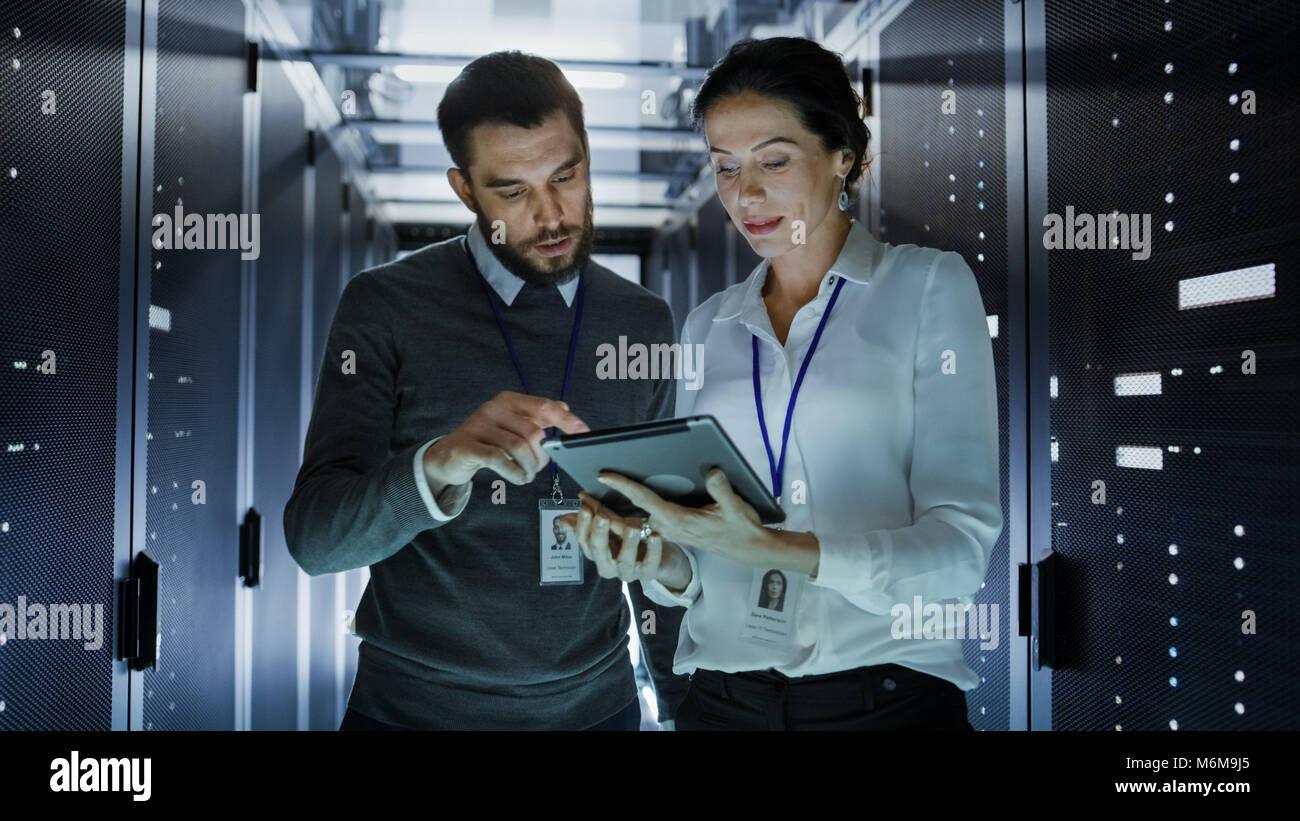 Maschio e femmina di tecnici IT nel Data Center / sala server, essi stanno parlando. Uno di essi detiene il computer Tablet. Foto Stock