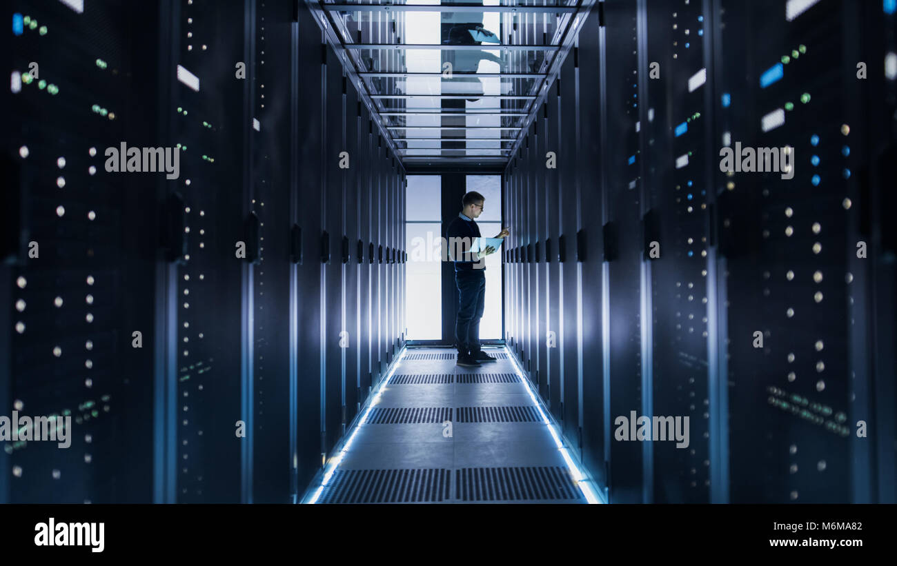 Maschio Ingegnere IT funziona su un computer portatile alla fine di un corridoio in un grande centro dati. Righe di server rack sono visti. Foto Stock
