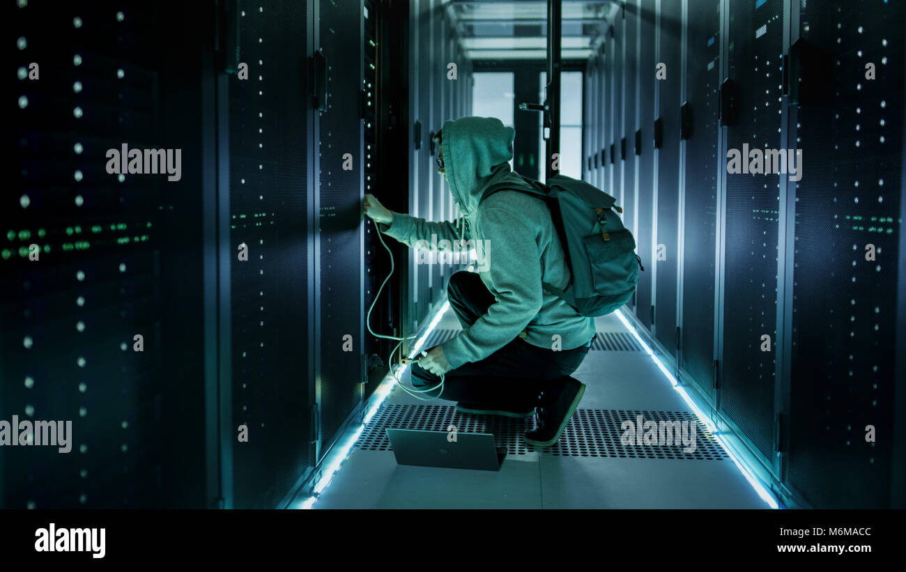 Un Hacker incappucciato con il computer portatile si connette al server in rack e ruba le informazioni dai data center aziendali. Foto Stock