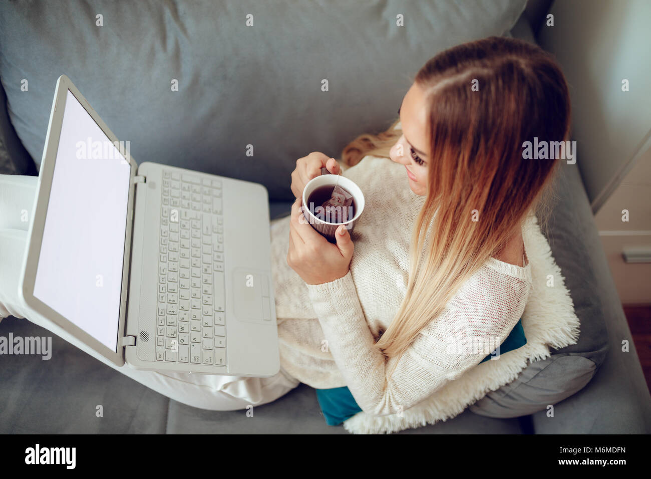 Attraente ragazza seduta al suo soggiorno con tazza di tè e utilizzando laptop. Ella è la navigazione in rete e del controllo dei mezzi di comunicazione sociale. Vista dall'alto. Foto Stock