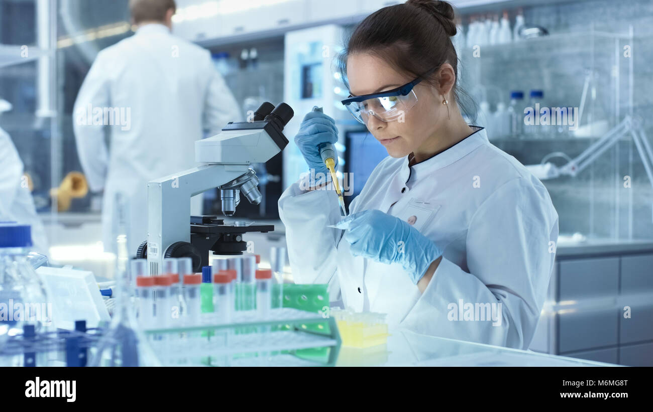 Ricerca di donne scienziato utilizza micropipetta riempire le provette per la prova in un grande e moderno laboratorio. Sullo sfondo gli scienziati stanno lavorando. Foto Stock