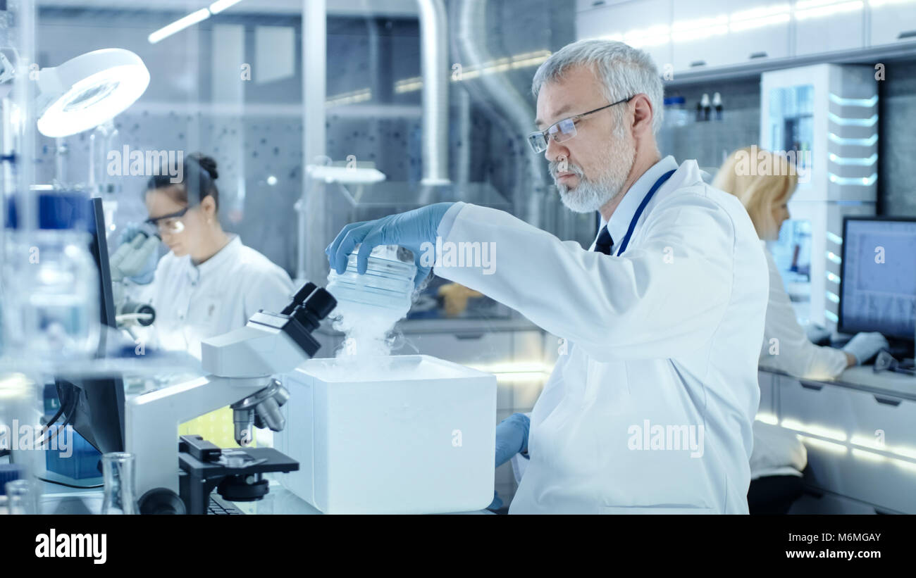 Senior Medical Research Scientist prende su piastra di Petri con campioni dal frigorifero aperto Box. Egli lavora in un affollato laboratorio moderno Centro. Foto Stock