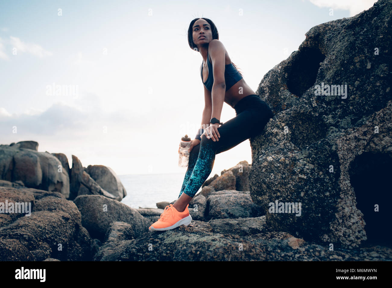 Un sano donna africana con una bottiglia di acqua seduto su una roccia. Rilassante femmina dopo esercizio all'esterno. Foto Stock