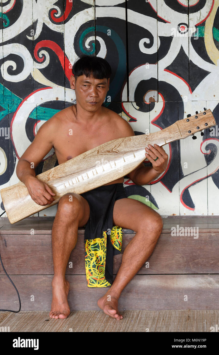 Uomo nativa la riproduzione di musica tradizionale con un elettrico Sape in Orang Ulu Longhouse, prima di albero della vita murale nel Sarawak Villaggio Culturale Foto Stock