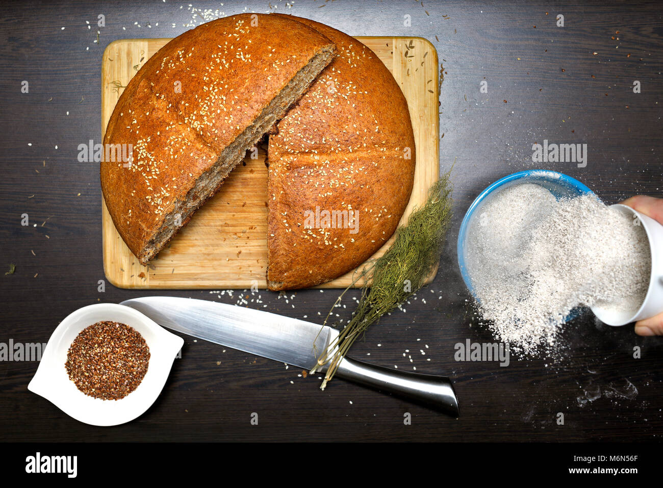 Fatto in casa pane integrale con semi di lino, farina integrale, aneto e coltello su un tavolo di legno. Donna diffondendo la farina in una ciotola. Foto Stock