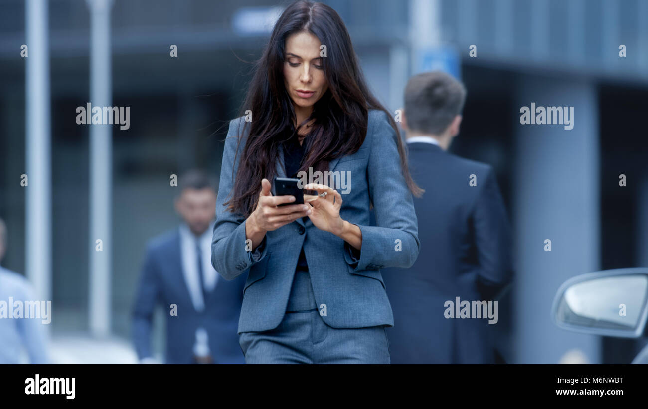 Business donna in abito realizzato su misura camminando sulla trafficata città grande Street nel quartiere degli affari, controlli il suo smartphone. Fiducioso donna sulla strada. Foto Stock