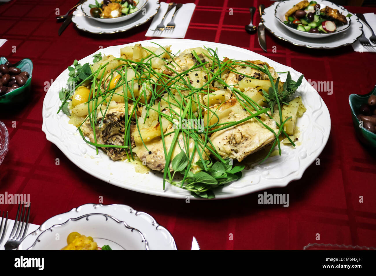 Greco aglio limone pollo e patate servita del piatto bianco sulla tovaglia rossa circondata da ciotole di insalata greca e una tazza di olive kalamata Foto Stock
