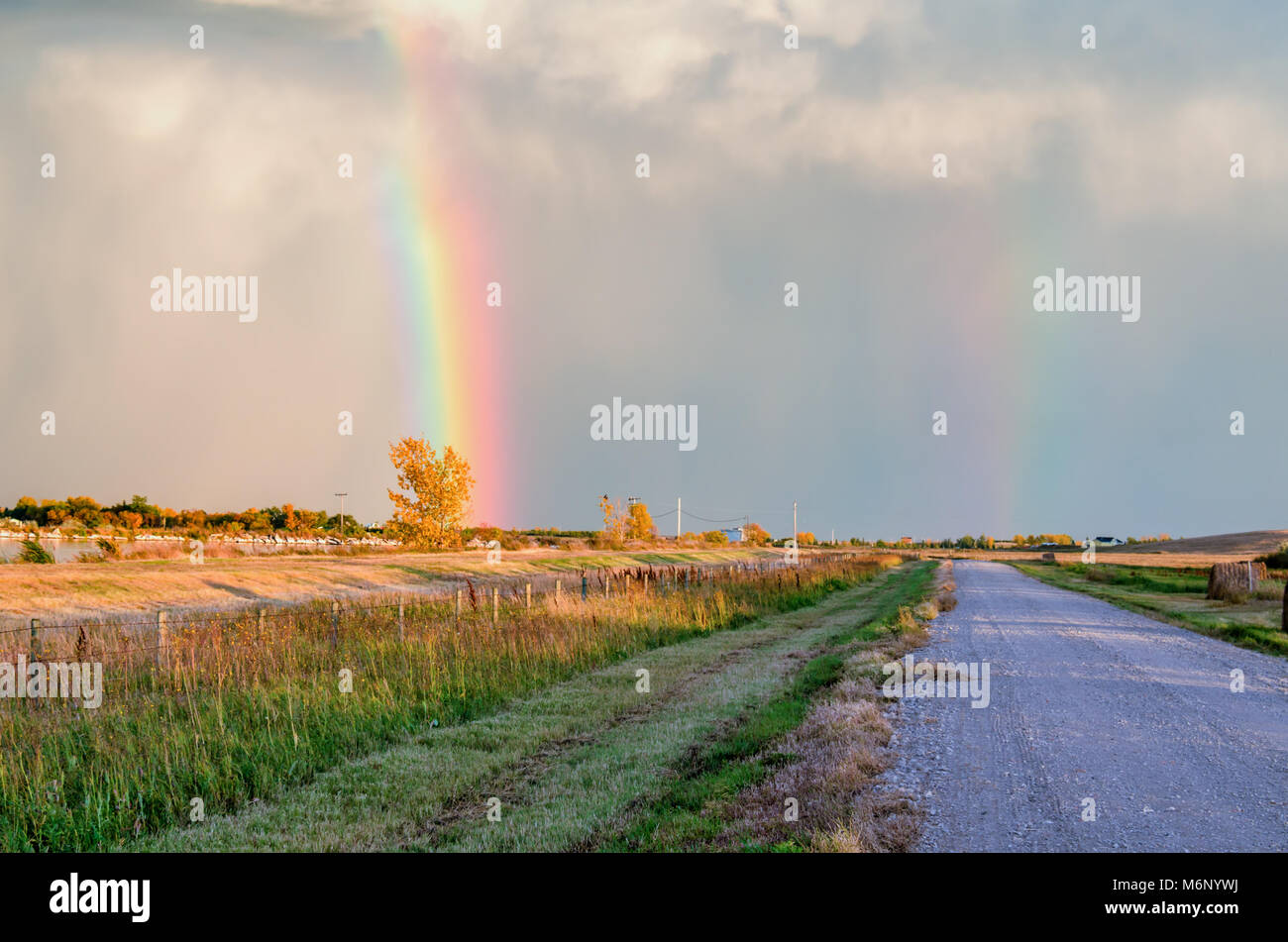 Una strada sterrata lungo il lago e i campi con haystacks alla fattoria, blu grigio nuvole e un multicolore doppio arcobaleno nel cielo. Foto Stock