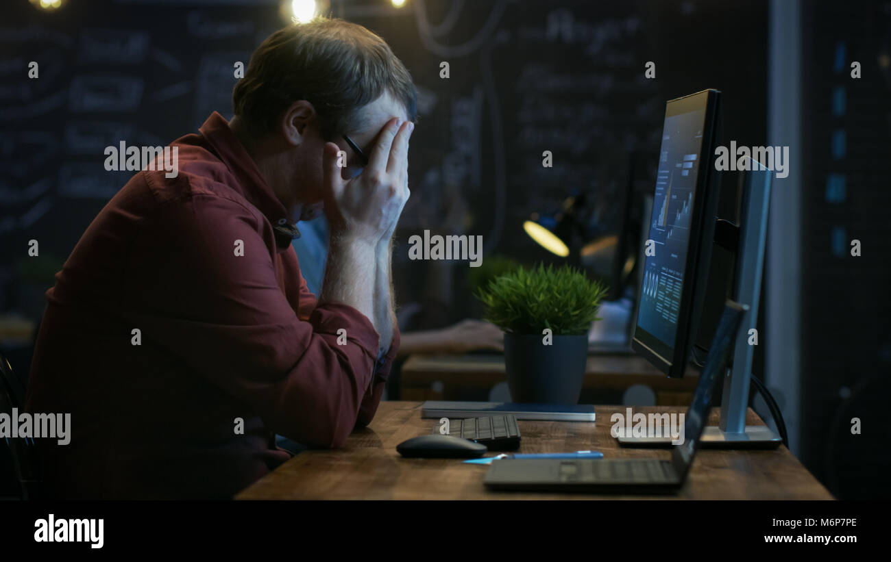 Sottolineato finanziere colpisce la tavola con il pugno di frustrazione e copre il volto con le mani. Sta lavorando su un Personal Computer con le statistiche Foto Stock